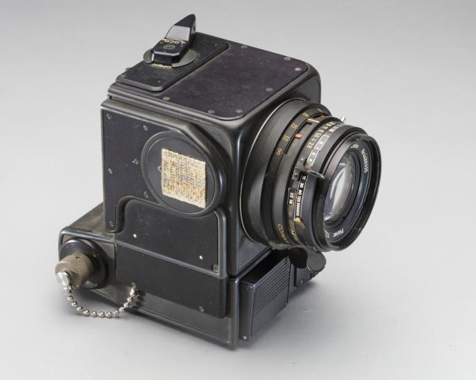 A black, box-shaped camera with 70mm lens and piece of velcro attached to side above silver chain leading to plug