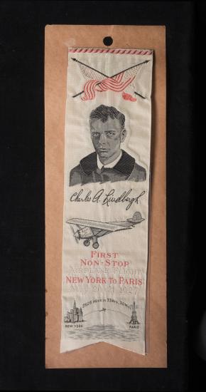 A white ribbon with two flags over black and white image of Charles Lindbergh. Below Charles Lindbergh is a commemoration of the first non-stop airplane flight between New York and Paris.