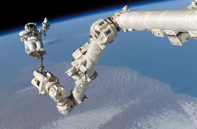 An astronaut performs work outside of the International Space Station using a special arm-shaped object. He is attached to the object by only a foot restraint.