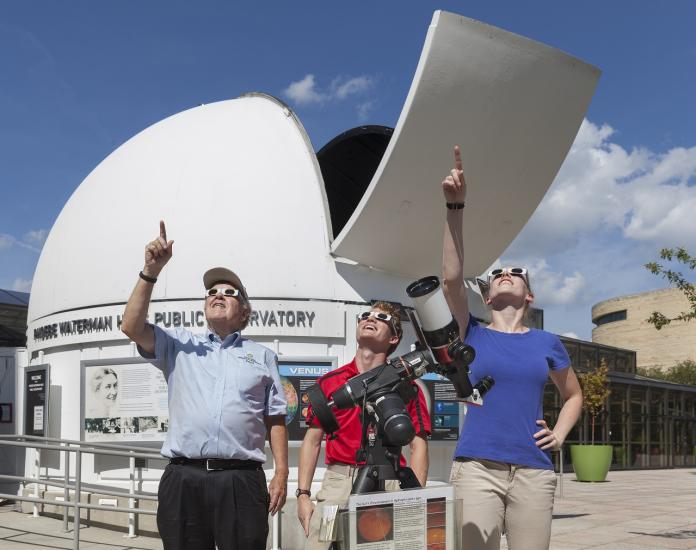 Three people observing a solar eclipse with safe solar glasses and a filtered telescope.