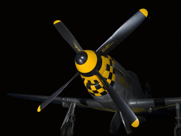 Four-blade propellers on nose of gray and yellow checkered P-51 Mustang aircraft