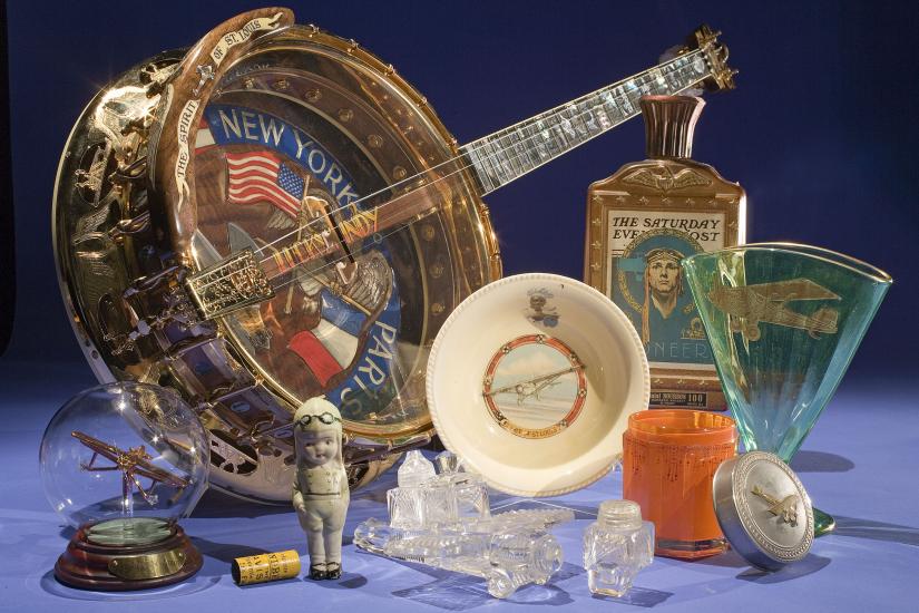 An assorted set of items commemorating Charles Lindbergh including a banjo, a bottle of alcohol, a glass ornament, and a white bowl.