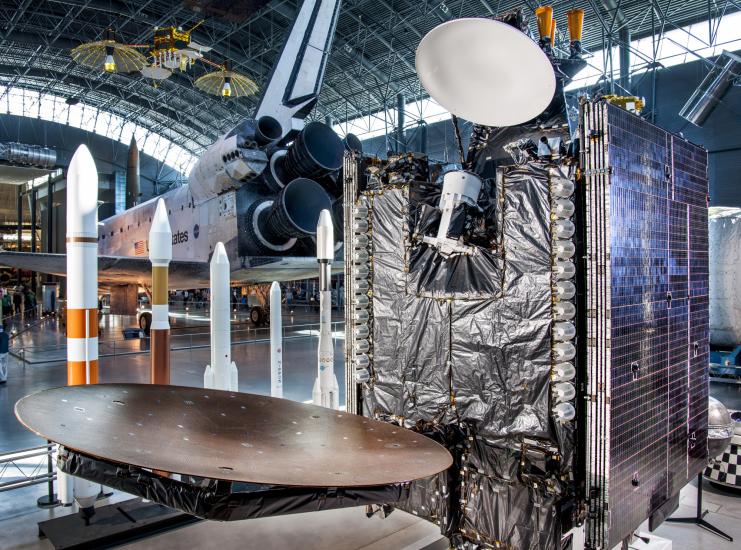 Partial view of backup communications satellite on display at the Museum. The base features a cube-like shape with an antenna dish hanging from the visible side and a second antenna dish hanging from top of the base. Two sides feature solar panels whereas the other two visible sides feature black wrap.