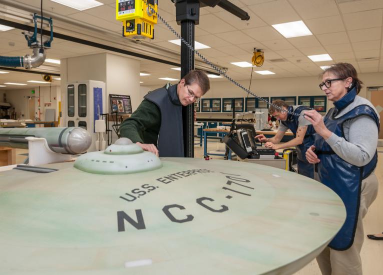 Three Smithsonian staff members use an X-ray machine to take images of the interior of a dish-shaped spaceship studio model.