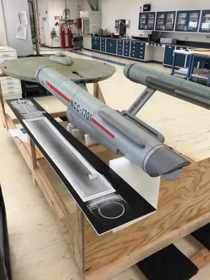 The Starship Enterprise, a gray-colored disc-shaped starship studio model with two rear, cylinder-shaped sections, is placed above an X-ray scan print of one of the rear sections of the model.