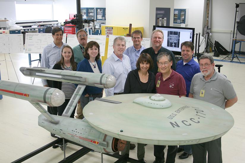 A group of Museum employees in charge of handling a gray spaceship studio model from the Star Trek series stand behind the studio model.