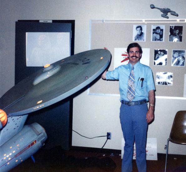 A white male poses next to a gray spaceship studio model from Star Trek.