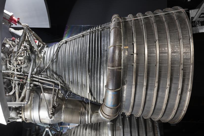 Side view of a rocket engine on display in the museum. One large, conic-shaped nozzle is attached to a fuel manifold pipe at the center of the nozzle. Cooling tubes are attached throughout the nozzle.