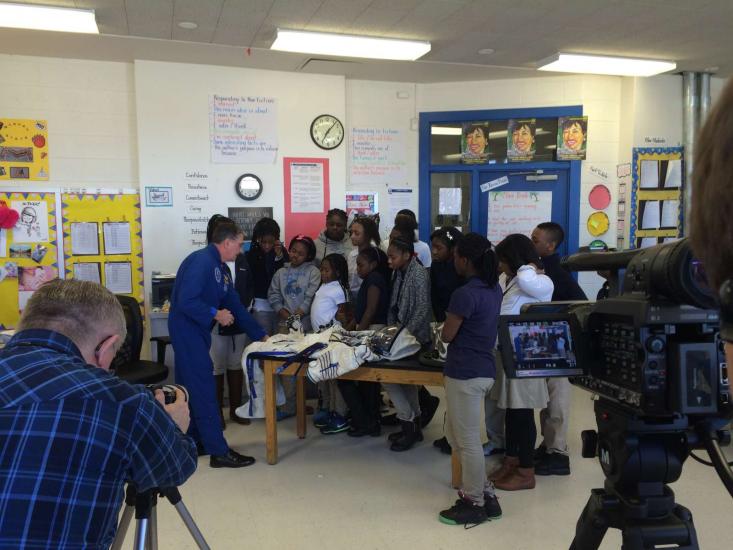 An astronaut wearing a blue full-body suit speaks to several students inside a classroom. Multiple camerapeople are filming the interaction.