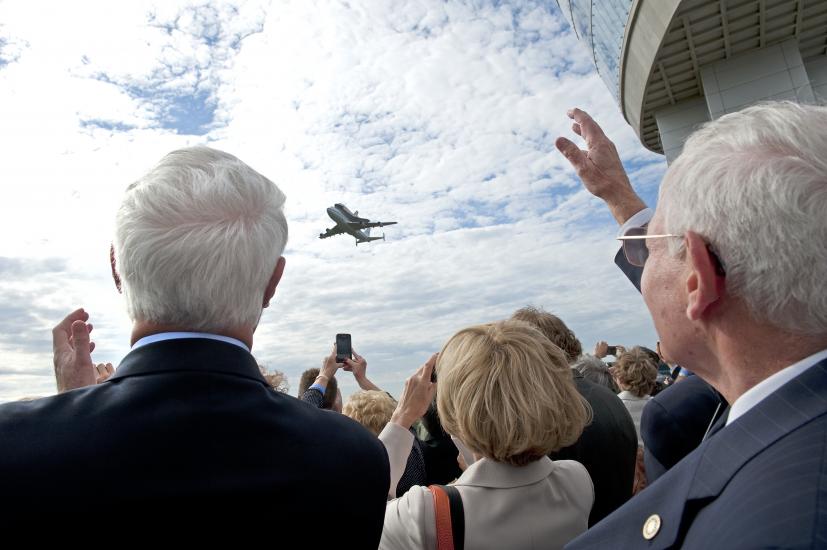 A group of people watch as the Space Shuttle Discovery, attached to a larger Boeing aircraft, flies low to the ground above the Steven F. Udvar-Hazy Center.