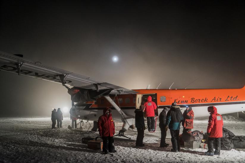 A group of people stand near a orange and white monoplane in Antarctica.