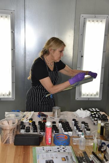 Smith holds up a board that she is painting with different colors. In front of her is a workbench with all of her paints. 