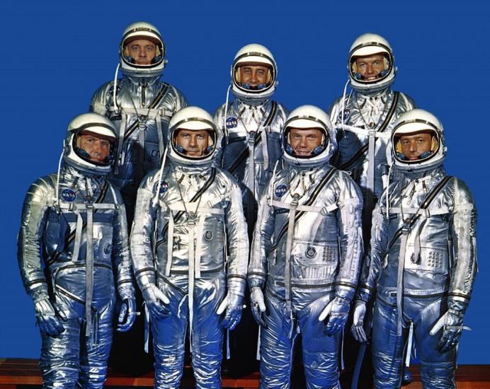 These seven individuals composed the first group of astronauts announced by the National Aeronautics and Space Administration. They stand in shiny metallic suits and smile at the camera.