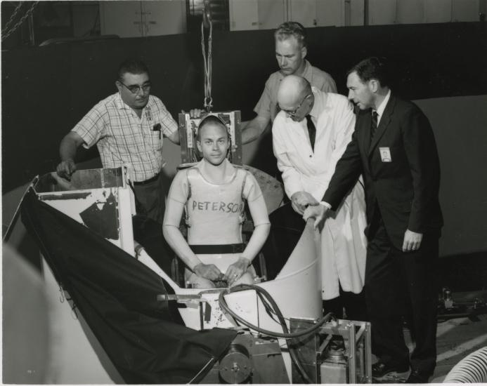 Man sits inside a centrifuge pod, four others surround him. 