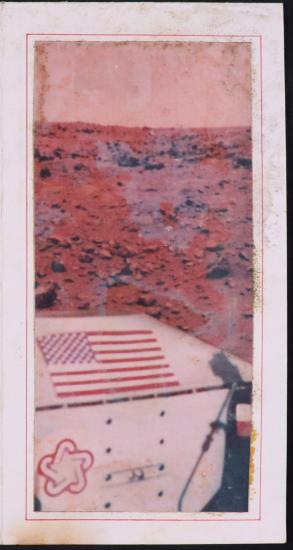 foreground: close up of Viking lander with American flag and 1976 bicentennial symbol (white five-point star inside a stylized star of red, white and blue). Background: red rocky ground of Mars