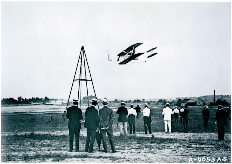 Black and white photo of the Wright Flyer in flight with crowd of onlookers below. 