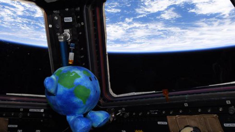 Celestial Buddies plush Earth toy is floating in the international space station. It is shaped and colored like a small Earth with a suprised expression. It is looking out a window towards Earth. 