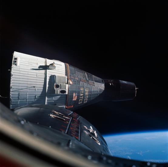 View of the Gemini VII spacecraft, a white and dark-gray colored conic object, from a window inside the Gemini VI spacecraft.