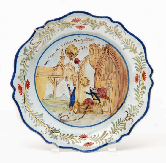 Ceramic plate with painted scene of two people preparing balloon basket for flight.