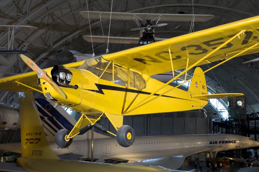 A Piper J-3, a yellow monoplane with one engine, is displayed from a suspended position inside the Museum.