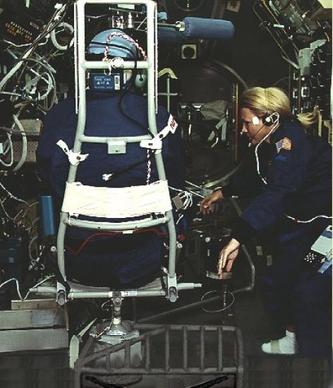 Astronaut on left sits in a chair with back turned while astronaut on right manages equipment.  