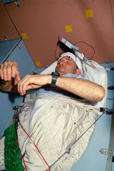 Astronaut strapped into their bed with white headband over their forehead.