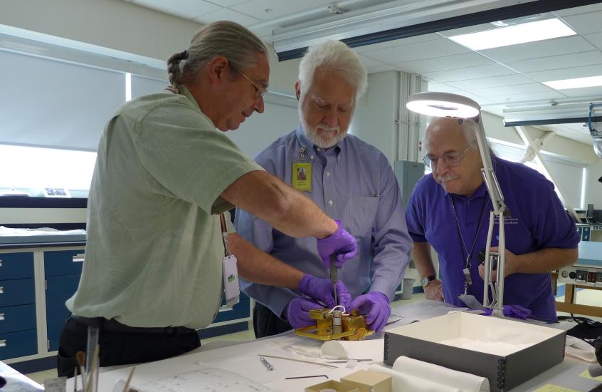 Three people stand around the film transport and work on it on a table. 