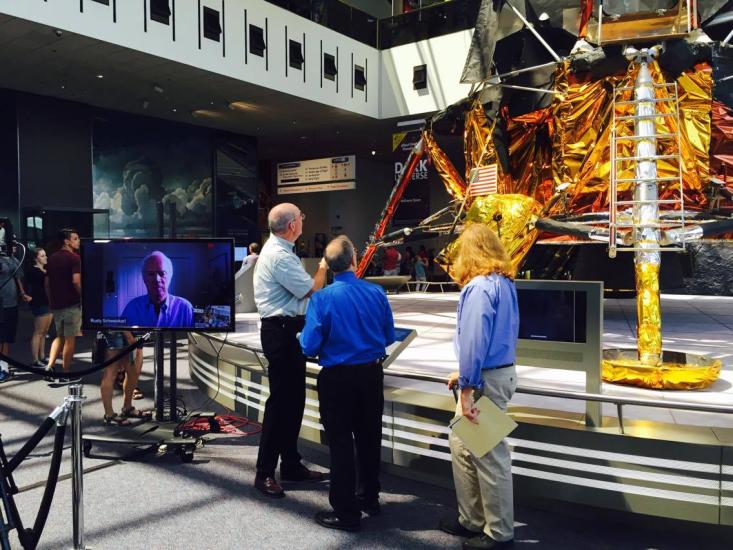 Three speakers with backs to camera look at the lunar module. Next to them is a TV screen with guest astronaut. 