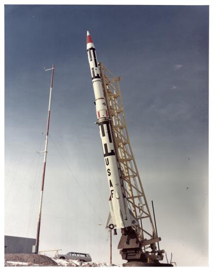 White rocket with red and black details, USAF painted on side, on yellow scaffolding.  Car and red and white pole in the background