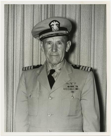 Half-length portrait of man in light colored summer uniform with hat, posed standing in front of a light-colored curtain