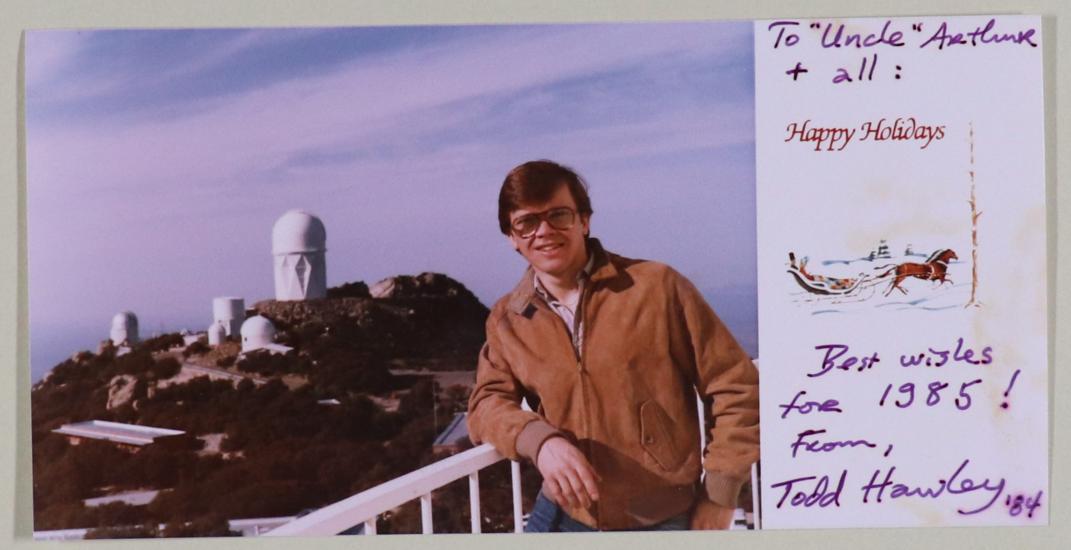 (left) Man with brown hair and glasses, wearing a tan jacket, stands at a railing. Background is an observatory with white buildings on a hill. (right) preprinted message "Happy Holidays" with drawing of horses and sleight. Handwritten note.