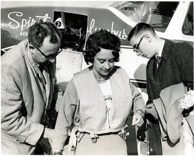 Black and White photograph: Airplane in the back has writing: Spirit of Columbus. Woman in the center has dark hair and wears and inflatable vest. Men on left and right wear glasses and are turning towards the woman.