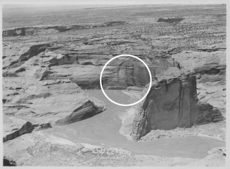 A black and white photograph showing large vertical rock formations that house cliff dwellings. The dwelling area is circled by a superimposed white circle.