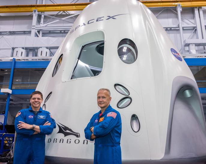 two astronauts in blue flight suits in from of capsule