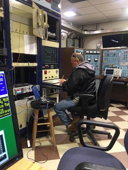 Man works at a small desk surrounded by equipment. 