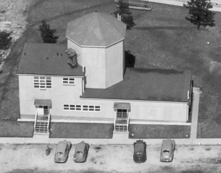 Aerial photo of a building with one large silo and cars parked out front. 
