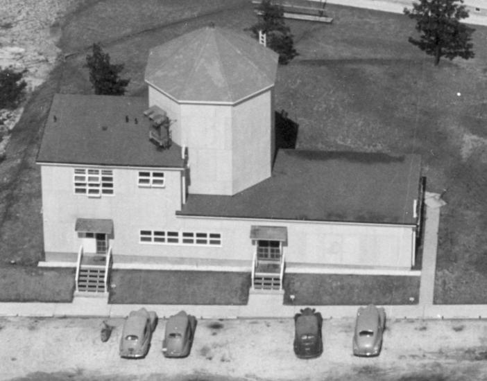 Aerial photo of a building with one large silo and cars parked out front. 