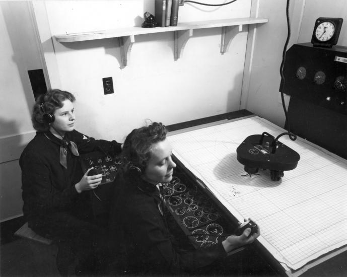 Two women sit in front of controls and plotting table. 