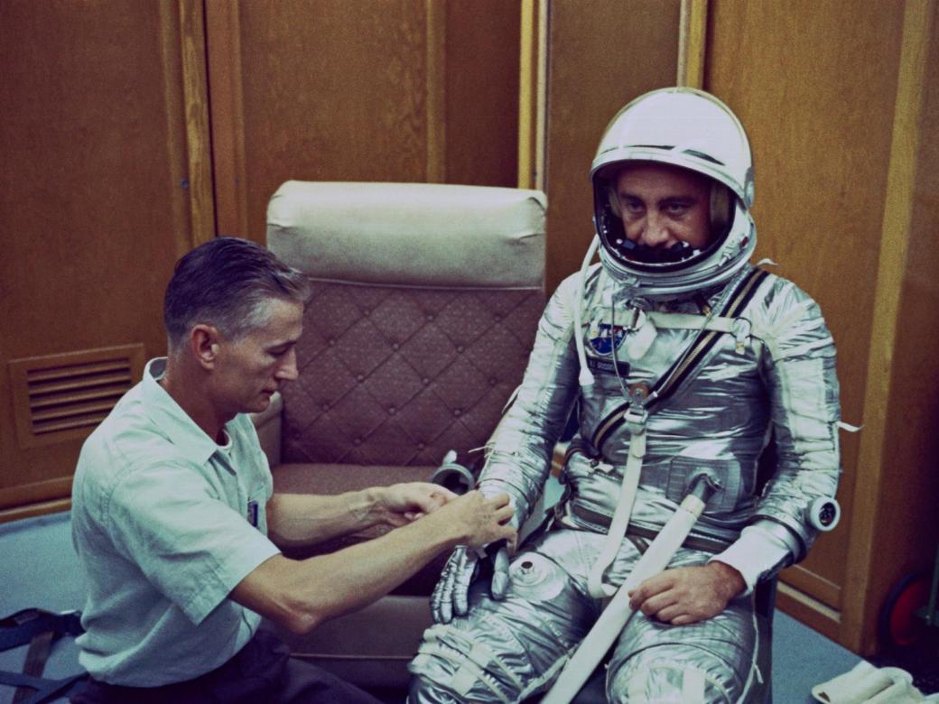 man in silver spacesuit with another man assists him with his glove