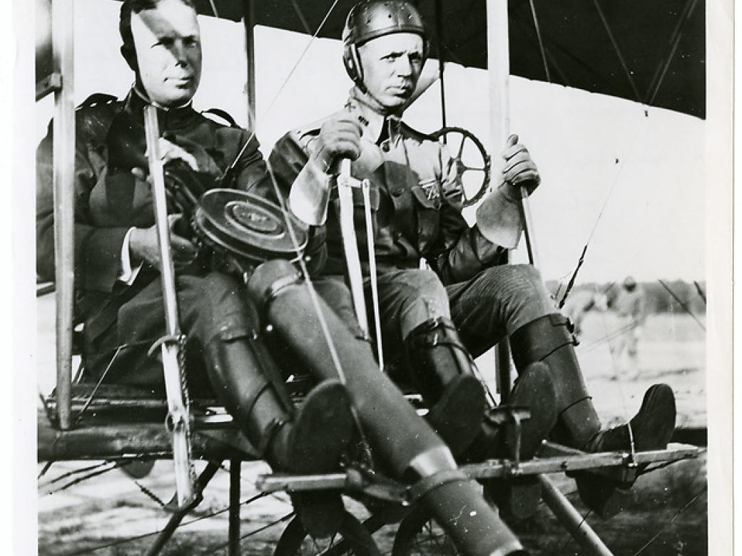 Two men in the seat of a Wright airplane. The man on the left is holding a large machine gun and the man on the right is at the controls.