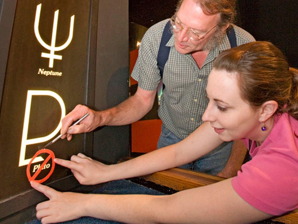 Two people place a red circle with a slash through it over the word "pluto" outside an exhibit entrance.