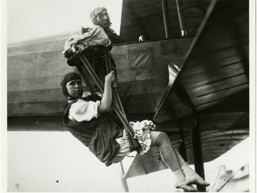 A woman hangs from behind the wing of an airplane, looking towards the camera. A man sits in the cockpit, also looking toward the camera.