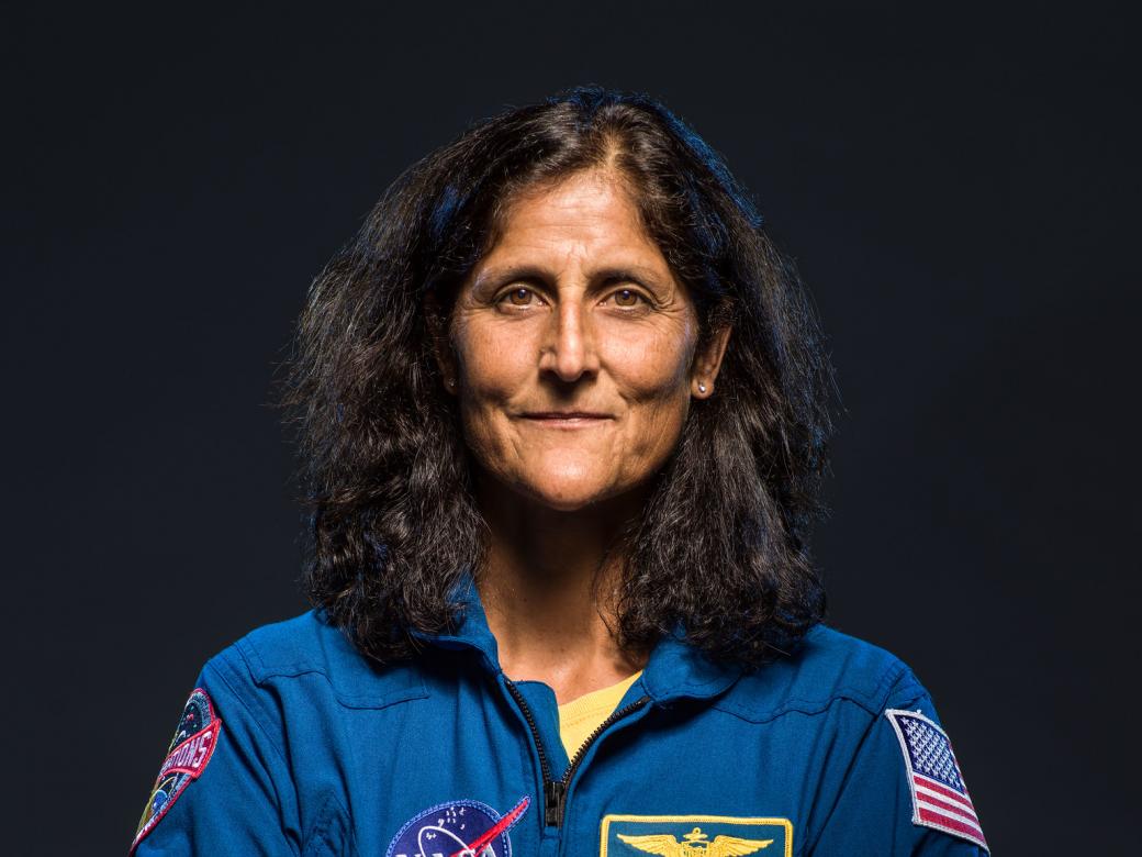 NASA Astronaut Sunita Williams looks at the camera, with a dark background, in a blue suit. 
